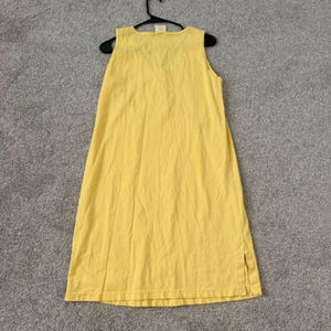 K&ouml;nnte beinhalten: Ein &auml;rmelloses, gelbes Kleid h&auml;ngt an einem schwarzen Kleiderb&uuml;gel. Das Kleid ist aus einem weichen Stoff gefertigt und hat ein schlichtes, l&auml;ssiges Design. Das Kleid ist knielang und hat einen Schlitz an der Seite.