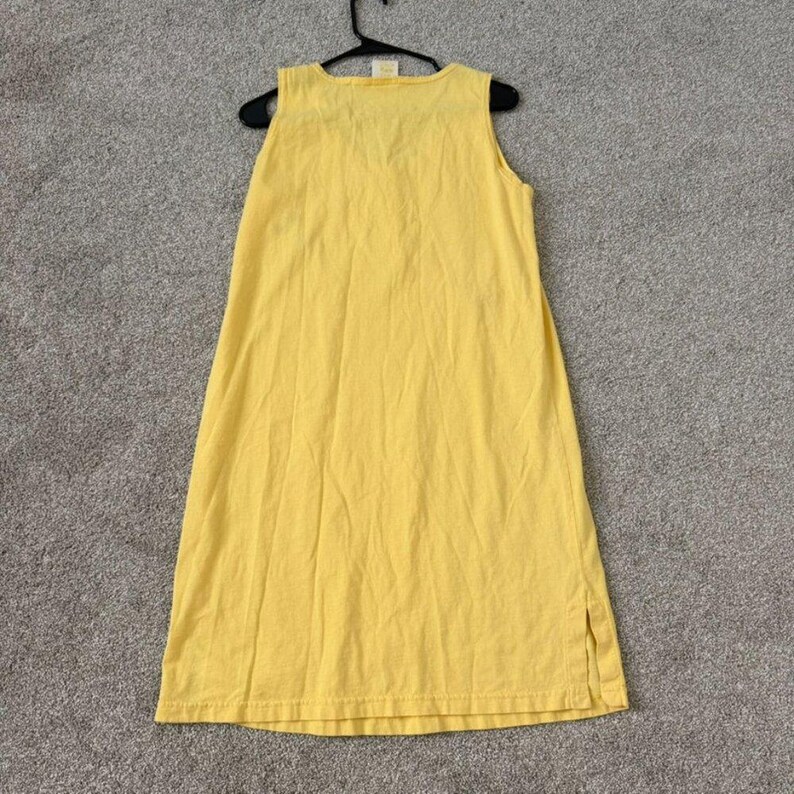 K&ouml;nnte beinhalten: Ein &auml;rmelloses, gelbes Kleid h&auml;ngt an einem schwarzen Kleiderb&uuml;gel. Das Kleid ist aus einem weichen Stoff gefertigt und hat ein schlichtes, l&auml;ssiges Design. Das Kleid ist knielang und hat einen Schlitz an der Seite.