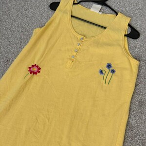 K&ouml;nnte beinhalten: Gelbes &auml;rmelloses Kleid mit gestickten floralen Details. Das Kleid hat einen Rundhalsausschnitt, eine Knopfleiste und gestickte Blumen in Rosa und Blau. Das Kleid h&auml;ngt an einem schwarzen Kleiderb&uuml;gel.