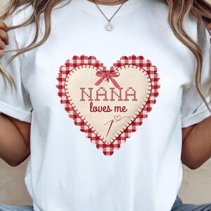 May include: White t-shirt with a heart-shaped appliqué. The heart has a red gingham border and the words "NANA loves me" in a stitched font. A small bow and needle illustration are also present.