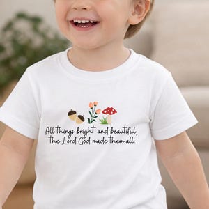 Op de afbeelding: Wit T-shirt met een ontwerp van eikels, bloemen en een paddenstoel. De tekst op het shirt luidt: "All things bright and beautiful, The Lord God made them all."