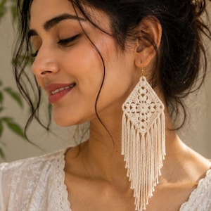 May include: A long, cream-colored crochet earring featuring a diamond-shaped motif with intricate floral-like patterns. The lower portion of the earring consists of numerous thin, hanging fringe threads that taper to a point at the bottom.