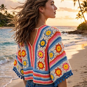 May include: A crochet top featuring a vibrant granny square pattern with yellow, orange, and pink floral motifs on a cream background, accented by horizontal stripes in blue, pink, and orange, finished with blue pom-pom trim on the sleeves.