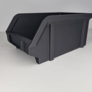 May include: A black plastic storage bin with a slanted front and a rectangular shape. The bin has a lip on the front and sides, and is designed for easy stacking. The bin is made of a durable plastic material.