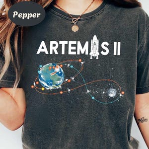 May include: Dark gray t-shirt with the text "ARTEMIS II" in white. The shirt features a graphic of Earth, a rocket, and a depiction of a space mission's trajectory. The design includes stars and a moon, creating a space exploration theme.