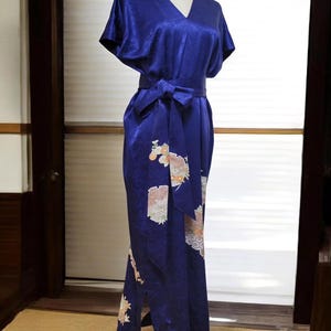 以下が含まれることがあります： Vネックと短い袖が付いた、ロイヤルブルーの着物風ドレス。 ドレスはウエストに結ばれたお揃いのベルトと、下部に花柄模様が特徴です。 ドレスはマネキンに飾られています。