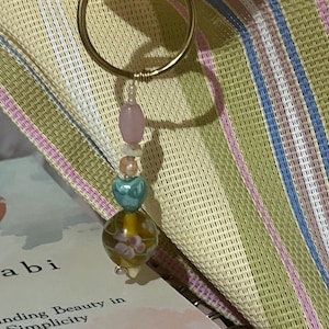 Puede incluir: Un pendiente de aro dorado con un dije colgante. El dije presenta una cuenta rosa, un grupo de pequeñas cuentas blancas, una cuenta en forma de corazón turquesa y una cuenta grande, redonda, amarillo-marrón con detalles florales. Sobre un fondo a rayas.