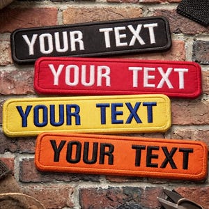 Könnte beinhalten: Vier rechteckige, gestickte Aufnäher in Schwarz, Rot, Gelb und Orange, jeweils mit dem Text "YOUR TEXT" in kontrastierenden Farben. Die Aufnäher werden vor einem Backsteinmauer-Hintergrund gezeigt.