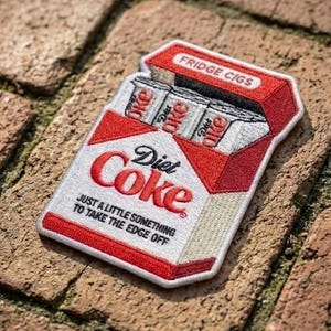 May include: Embroidered patch in the shape of a Diet Coke cigarette pack. The patch is white with red accents and features the Diet Coke logo and the phrase "JUST A LITTLE SOMETHING TO TAKE THE EDGE OFF." The top of the pack reads "FRIDGE CIGS."