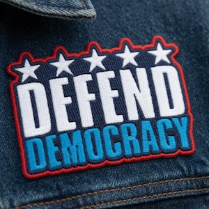 Puede incluir: Primer plano de una chaqueta vaquera con un parche que dice "DEFEND DEMOCRACY". El parche presenta texto blanco sobre un fondo azul marino, delineado en rojo y azul, con cinco estrellas blancas en la parte superior.