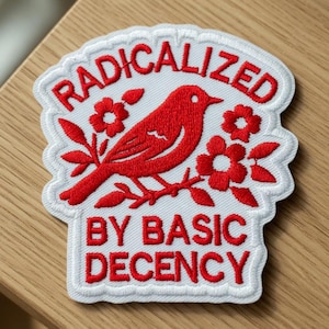Puede incluir: Parche bordado con un pájaro rojo posado en una rama florecida, rodeado de las palabras "RADICALIZED" y "BY BASIC DECENCY" en letras rojas sobre un fondo blanco. El parche tiene un borde blanco.
