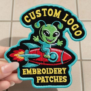 Puede incluir: Parche bordado con un alienígena verde montado en un cohete rojo. El alienígena tiene ojos grandes y lleva auriculares. El parche tiene las palabras "CUSTOM LOGO" y "EMBROIDERY PATCHES" en letras amarillas sobre un fondo negro y turquesa.