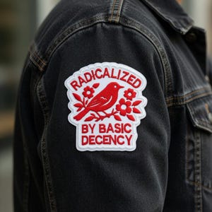 May include: A black denim jacket with a white and red embroidered patch. The patch features a red bird, flowers, and the words "RADICALIZED BY BASIC DECENCY" in red lettering.