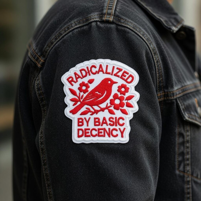 May include: A black denim jacket with a white and red embroidered patch. The patch features a red bird, flowers, and the words "RADICALIZED BY BASIC DECENCY" in red lettering.