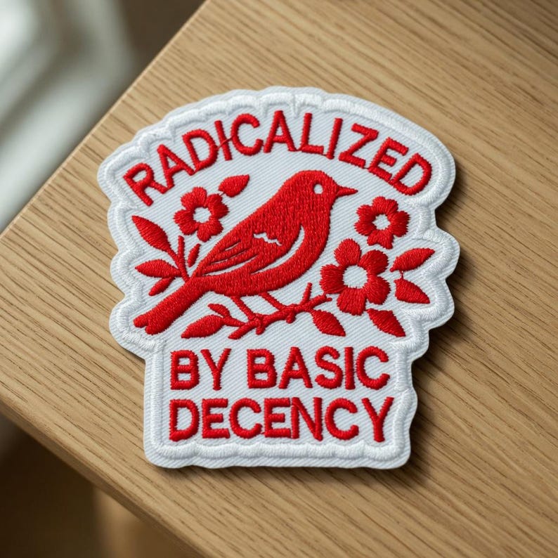 May include: Embroidered patch with a red bird perched on a flowering branch, surrounded by the words "RADICALIZED" and "BY BASIC DECENCY" in red lettering on a white background. The patch has a white border.