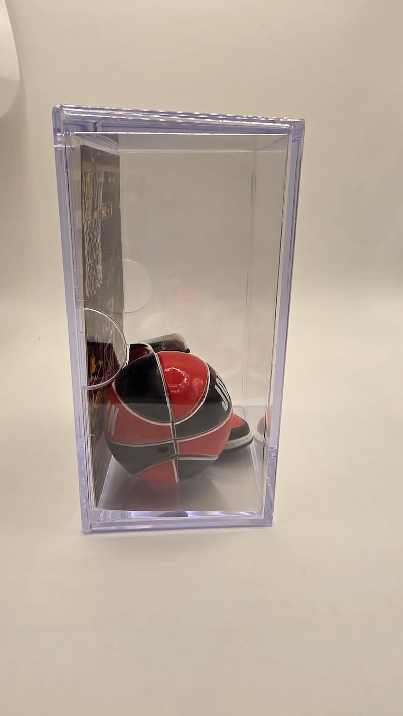 May include: A miniature basketball in a clear display case. The basketball is red, black, and white, with a segmented design. The case is rectangular and transparent, showcasing the collectible item.