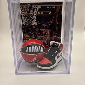May include: A clear display case containing a basketball and a miniature sneaker. The basketball is red with "JORDAN" in white lettering. The sneaker is black, white, and red. A basketball game image is in the background.