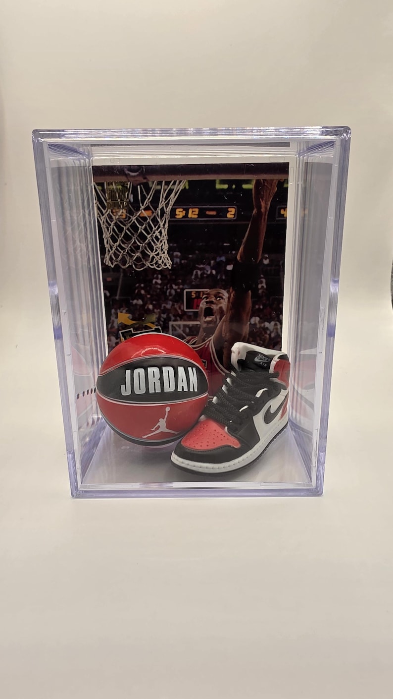 May include: A clear display case containing a basketball and a miniature sneaker. The basketball is red with "JORDAN" in white lettering. The sneaker is black, white, and red. A basketball game image is in the background.