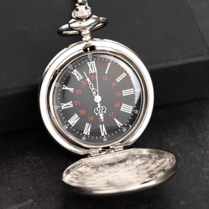 May include: A silver pocket watch with a black face and Roman numeral hour markers. The watch has a hinged cover that is open, revealing an engraved design. The watch is suspended by a chain.