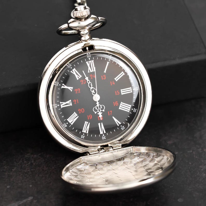 May include: A silver pocket watch with a black face and Roman numeral hour markers. The watch has a hinged cover that is open, revealing an engraved design. The watch is suspended by a chain.