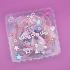 May include: A clear plastic case filled with pink and peach-colored hair extensions, decorative beads, and charms. The word "EDIKTED" is spelled out with colorful letter beads. The case is adorned with star, flower, and bow-shaped embellishments.
