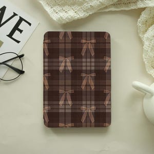 May include: A rectangular tablet case featuring a dark brown plaid pattern overlaid with a repeating design of small, light brown bows. The case has a smooth, flat surface and a consistent grid-like aesthetic across the entire front panel.