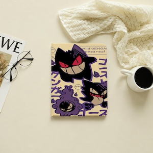 May include: A rectangular notebook with a light yellow cover featuring two purple Gengar characters, one with red eyes and a wide grin, the other with a tongue out. The text reads Wild GENGAR appeared! along with Japanese characters and icons.