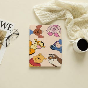 May include: A rectangular tablet case featuring a light beige background with a printed illustration of Winnie the Pooh, Tigger, Piglet, Eeyore, Roo, and Owl looking inward from the edges of the cover in a circular arrangement.