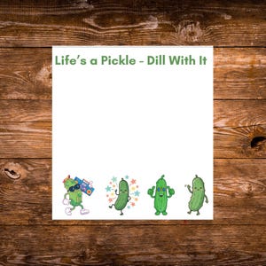 May include: A white notepad featuring the green text Life's a Pickle - Dill With It at the top. The bottom section displays four cartoon pickles: one with sunglasses and a boombox, one surrounded by stars, one with glasses giving thumbs up, and one waving.