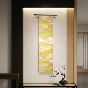 May include: A vertical wall hanging featuring a repeating golden bridge pattern on a light fabric, suspended from a dark horizontal rod. Below, a small bonsai tree with dark, twisted branches sits in a black pot on a low wooden platform.