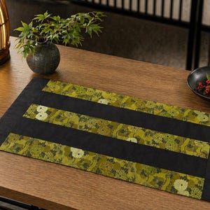 May include: A rectangular black and green floral patterned table runner. The runner has three horizontal floral patterned stripes in shades of green and white. The runner is on a wooden table.