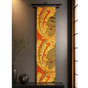 May include: A decorative Japanese wall hanging with a gold and orange design. The artwork features a repeating pattern of stylized floral and geometric shapes. The hanging is displayed on a black rod against a dark gray wall.