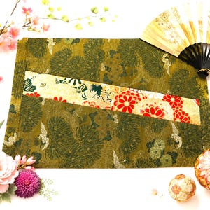 May include: A rectangular fabric mat featuring an olive green base with a repeating pattern of dark green pine needles and white cranes. A diagonal band across the centre displays a floral design with red, white, and gold accents.
