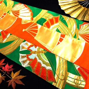 May include: An overhead shot of a colorful Japanese obi sash with a green and orange design. The sash features gold accents and fan motifs. A gold fan and autumn leaves are also visible.