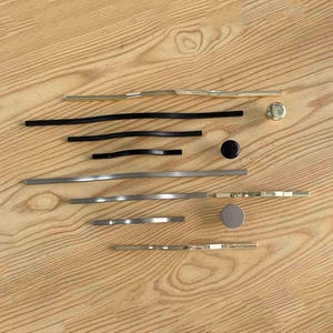 May include: A collection of cabinet hardware including various drawer pulls and knobs in gold, matte black, and silver finishes. The assortment features long, slender, slightly curved handles alongside circular knobs in matching metallic and black tones.