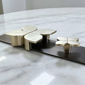 May include: Two brushed gold-toned metal cabinet knobs, each featuring a four-petaled flower shape. The larger knob has four distinct, rounded leaf-like segments, while the smaller knob is a compact version with a similar four-petal design.