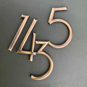 May include: Bronze metal house numbers 145 against a dark green background. The numbers are in a serif font and have a slightly weathered appearance, suggesting an antique or vintage style. The numbers are arranged in a visually appealing manner.