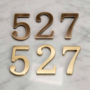 May include: Two sets of house numbers, 5, 2, and 7, arranged in two rows. The top row features numbers with a dark, brushed bronze finish, while the bottom row displays the same numbers in a lighter, polished brass finish with a metallic sheen.