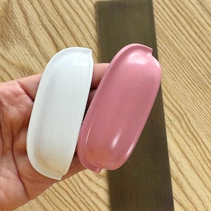 Op de afbeelding: Twee halfronde komgrepen, één in een matte witte afwerking en de andere in een zachtroze afwerking. Deze hardwarestukken hebben een glad, gebogen ontwerp met een verzonken greepgedeelte, bedoeld voor montage op kastdeuren of ladefronten.