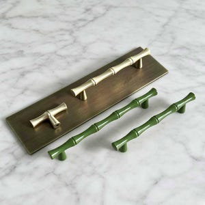 May include: A set of cabinet hardware featuring two long bamboo-style pulls and one small bamboo-style knob in a metallic gold finish, alongside two long green bamboo-style cabinet pulls. The hardware is displayed on a rectangular brass plate.