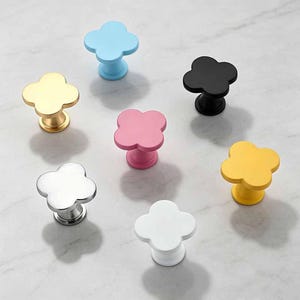 Puede incluir: Una colección de siete pomos para armarios con forma de trébol en varios colores, incluyendo azul claro, dorado, negro, rosa, plateado, amarillo y blanco. Cada pomo presenta un diseño de trébol de cuatro hojas con un poste central y superficie plana.