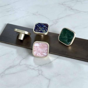 May include: Four square cabinet knobs with metallic gold-toned bases and smooth, rounded edges. The top surfaces feature distinct marbled patterns in blue, dark green, and light pink, with one knob shown from the side to reveal its mounting post.