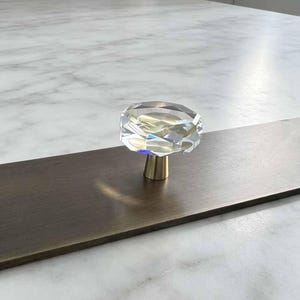 Könnte beinhalten: Ein Möbelknopf mit einem klaren, facettierten, rautenförmigen Glaskopf auf einem zylindrischen, messingfarben gebürsteten Metallsockel. Der Knopf ist auf einer flachen, rechteckigen, dunkelbronzefarbenen Metallplatte montiert.