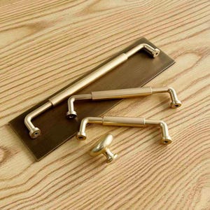 May include: A collection of brushed gold cabinet hardware including one long handle, two shorter handles, and one round knob, all arranged on a rectangular, dark bronze-toned metal display plate with visible screw holes at each end.