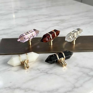 Puede incluir: Cinco pomos de armario de cristal de doble punta en rosa, rojo oscuro, transparente, blanco y negro. Cada pomo cuenta con una base de metal dorado y un alambre dorado envuelto alrededor del centro del cristal para su fijación en muebles.