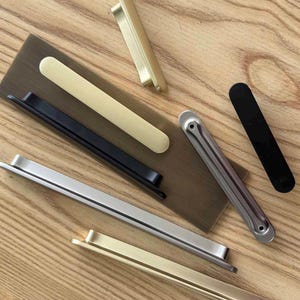 Op de afbeelding: Een collectie metalen kastgrepen in diverse afwerkingen, waaronder geborsteld goud, matzwart en zilver. De hardware-onderdelen hebben langwerpige, slanke en rechthoekige ontwerpen met afgeronde randen, sommige met schroefgaten voor montage.