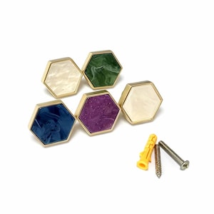 May include: Five hexagonal cabinet knobs with gold-toned metal frames and marbled acrylic inserts in white, green, blue, and purple. The set includes two white knobs, one green, one blue, and one purple, accompanied by two metal screws and one yellow anchor.