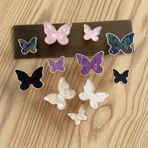 May include: A collection of butterfly-shaped knobs in various sizes and finishes, including iridescent blue, solid black, pearlescent white, marbled pink, and deep purple. Each knob features a gold-toned metal edge and a central decorative inlay.