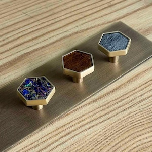 May include: Three hexagonal cabinet knobs with metallic gold-toned edges mounted on a rectangular brass plate. Each knob features a distinct inlay: one with iridescent blue and purple flecks, one with a textured brown surface, and one with grey.