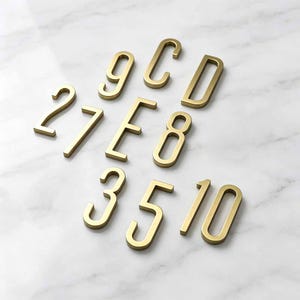 May include: A collection of gold-colored metal house numbers and letters including the characters 9, C, D, 2, 7, E, 8, 3, 5, 1, and 0. Each character features a clean, sans-serif font with a smooth, matte finish and a three-dimensional profile.